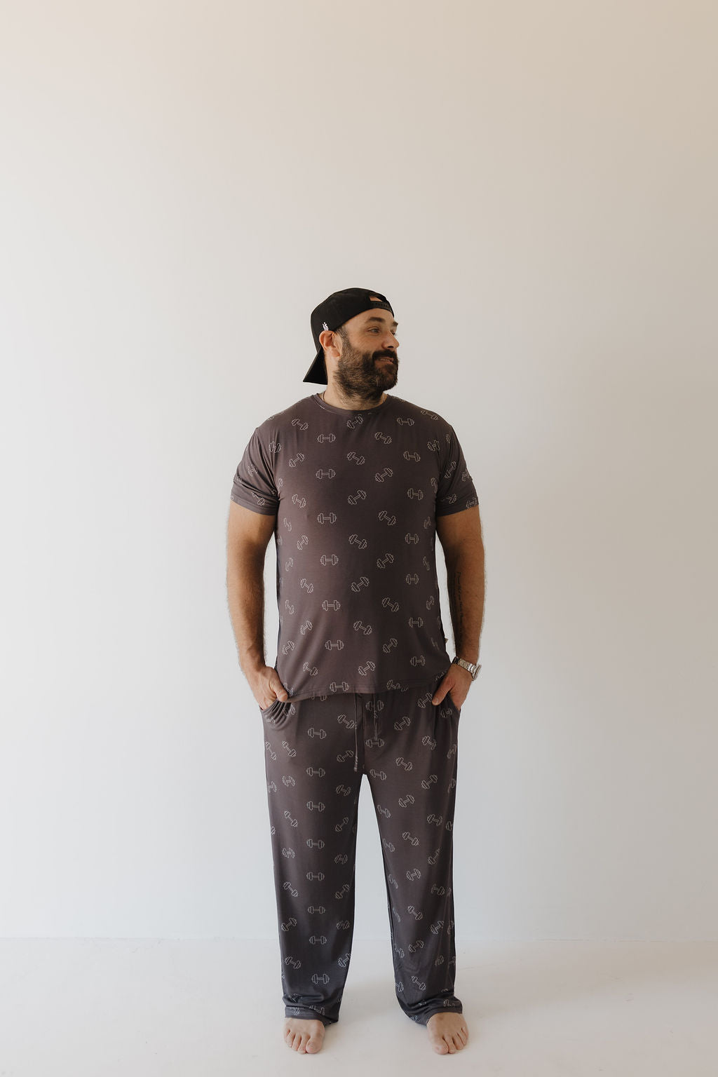 A bearded man stands barefoot against a white background, wearing the Bamboo Men's Short Sleeve Pajama in Charcoal Arm Day by forever french baby—a matching dark gray set with a subtle pattern. His hands are in his pockets as he looks to the side, finished with a black cap worn backward.