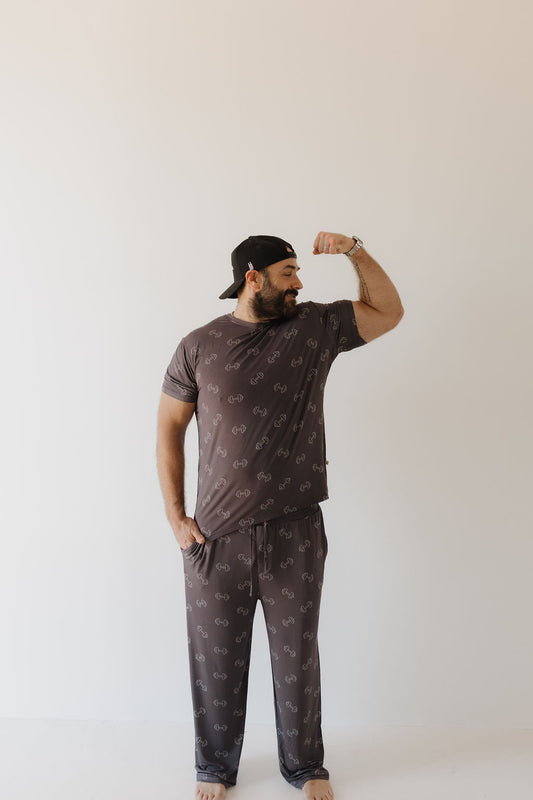 A man poses barefoot in hypoallergenic clothing from Forever French Baby, wearing the Bamboo Men's Short Sleeve Pajama set in Charcoal Arm Day. Sporting a backward black cap, he flexes his arm against a light backdrop.