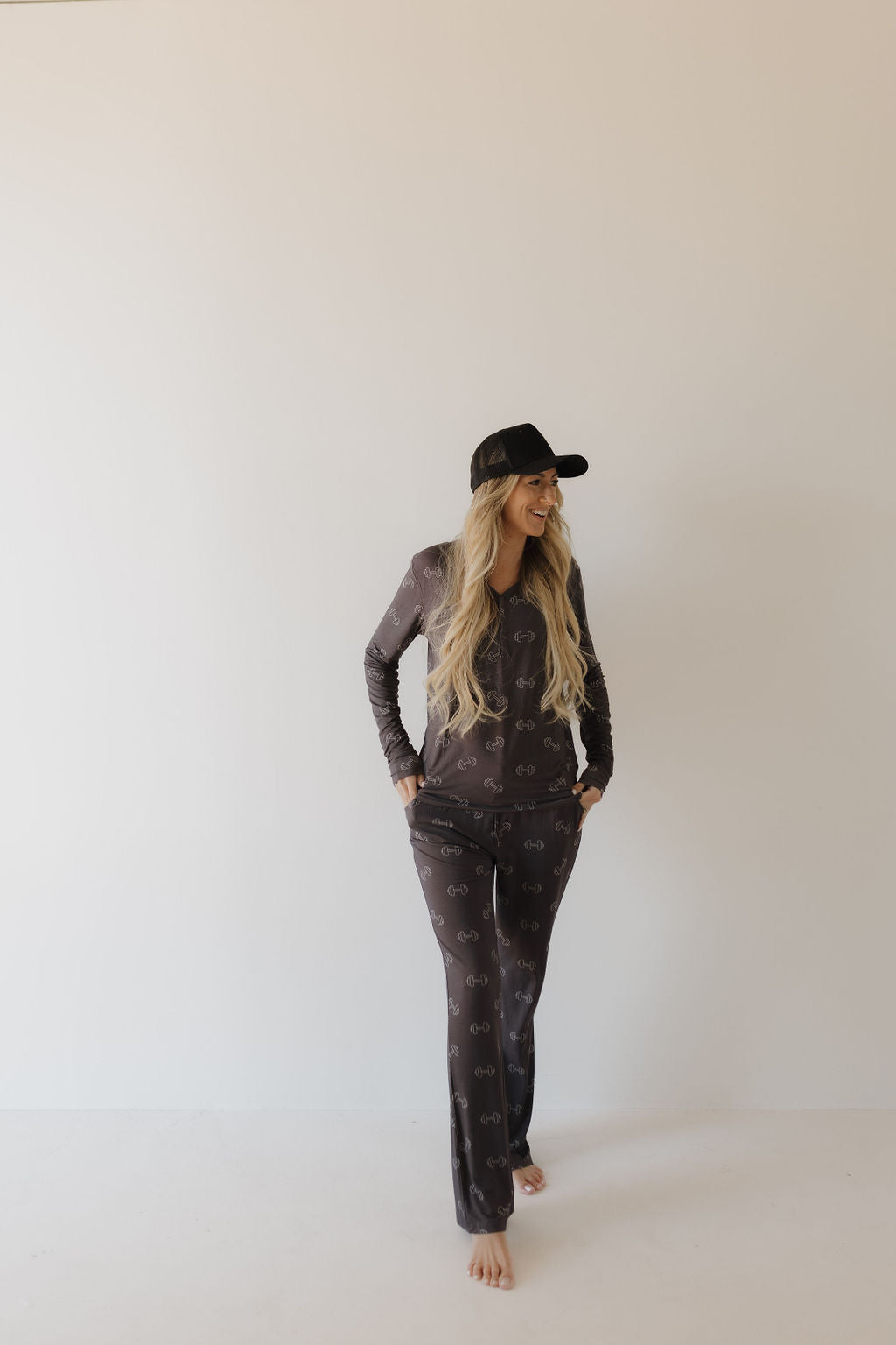 Barefoot against a plain backdrop, a person with long hair smiles warmly to the side, hands in pockets. They wear breathable clothing from forever french baby—a matching Women's Bamboo Pajama set named Charcoal Arm Day and a black cap.