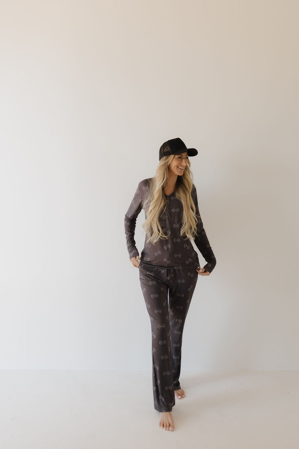 A person with long hair is wearing a black cap and breathable clothing from forever french baby, including Women's Bamboo Pajamas in Charcoal Arm Day with a subtle pattern, standing barefoot against a plain white background.