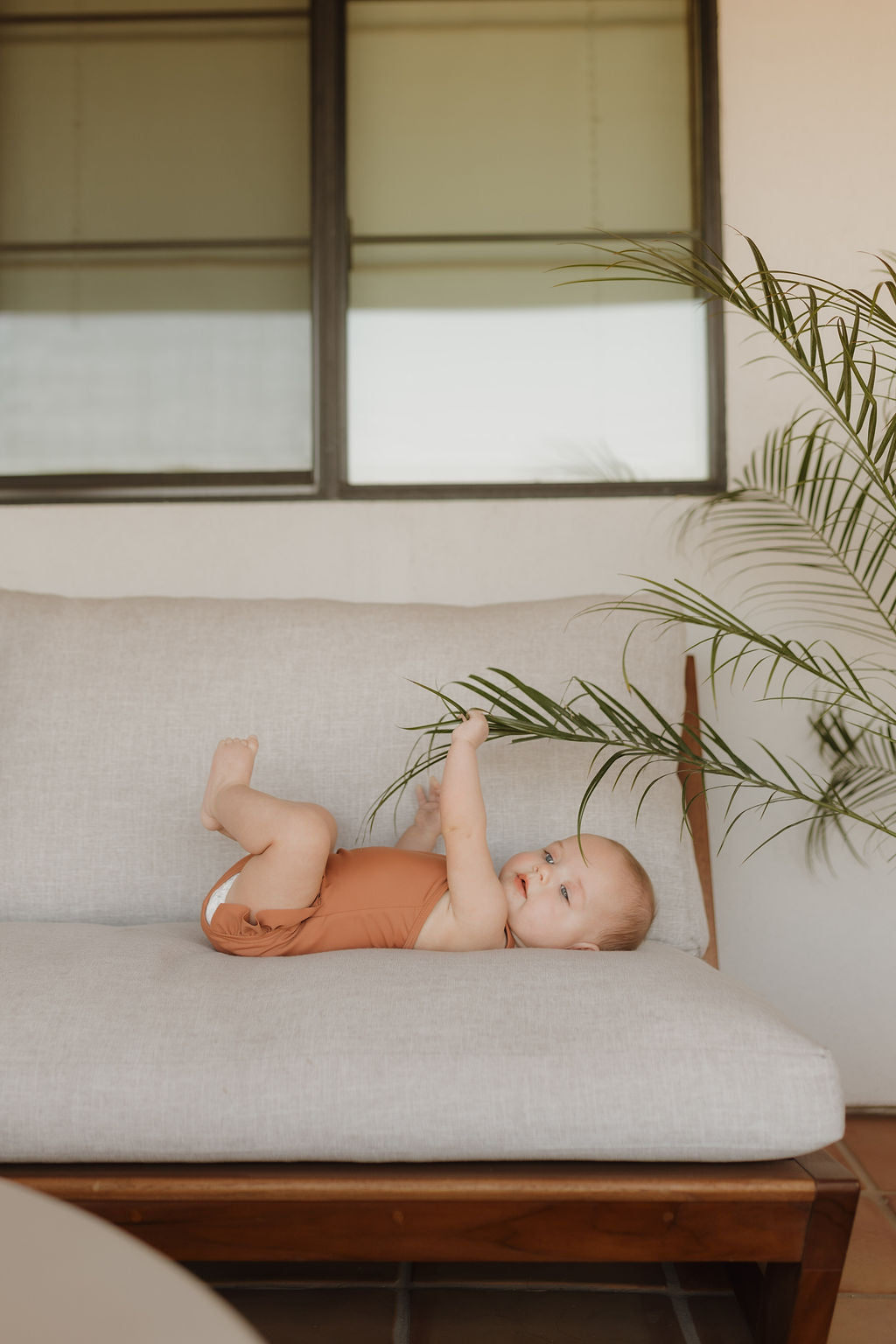 A baby in a brown romper from the Terra collection by forever french baby lounges on a beige sofa, engaging with a green palm leaf. A double-pane window softly lights the scene, embodying cozy elegance.