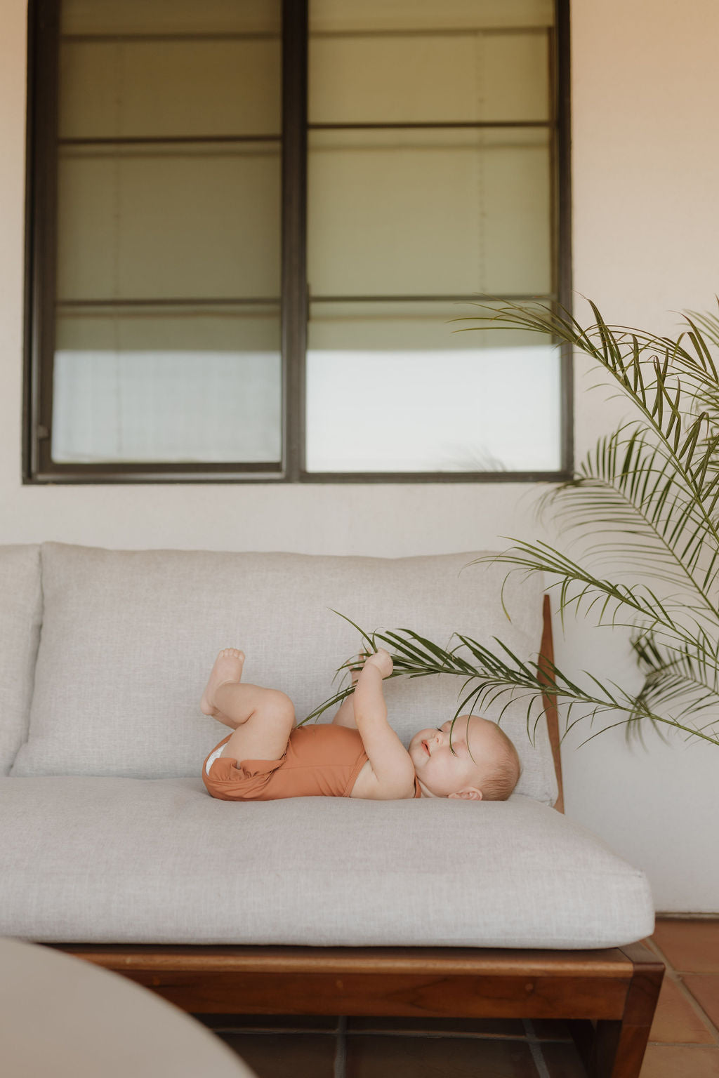 A baby from Forever French Baby's Terra collection, in an orange outfit, lies on a light gray couch, playfully holding a green palm frond under a large window.