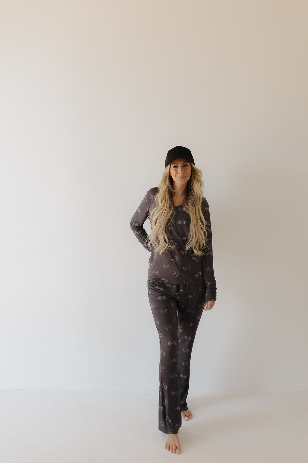 A woman with long blonde hair wears a black cap and dark outfit featuring Forever French Baby's Women's Bamboo Pajamas in Charcoal Arm Day. She stands barefoot against a plain white background, hand on hip, confidently looking at the camera in her stylish yet breathable attire.