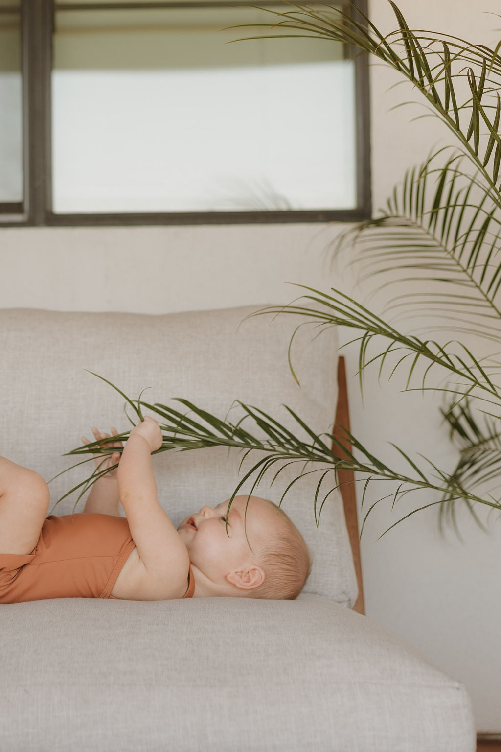 A baby in a vibrant orange Sleeveless Swimsuit from forever french baby's Terra collection lounges on a beige chair, cradling a palm leaf, with a window and lush greenery setting the scene for this charming moment.