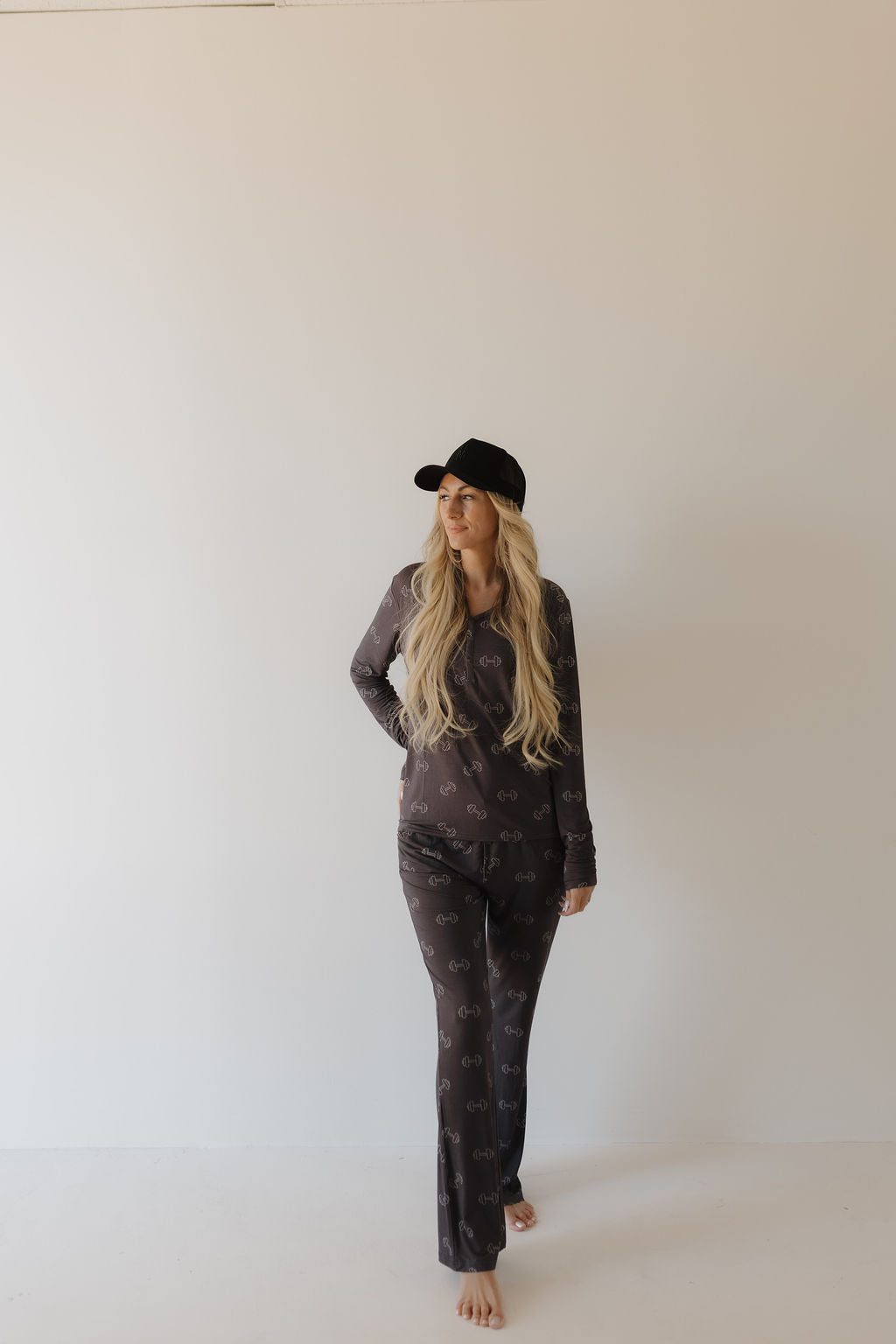 A woman with long blonde hair stands barefoot against a white background, wearing forever french baby's Women's Bamboo Pajamas in Charcoal Arm Day. She completes her look with a black hat and gazes to the right with a relaxed expression.