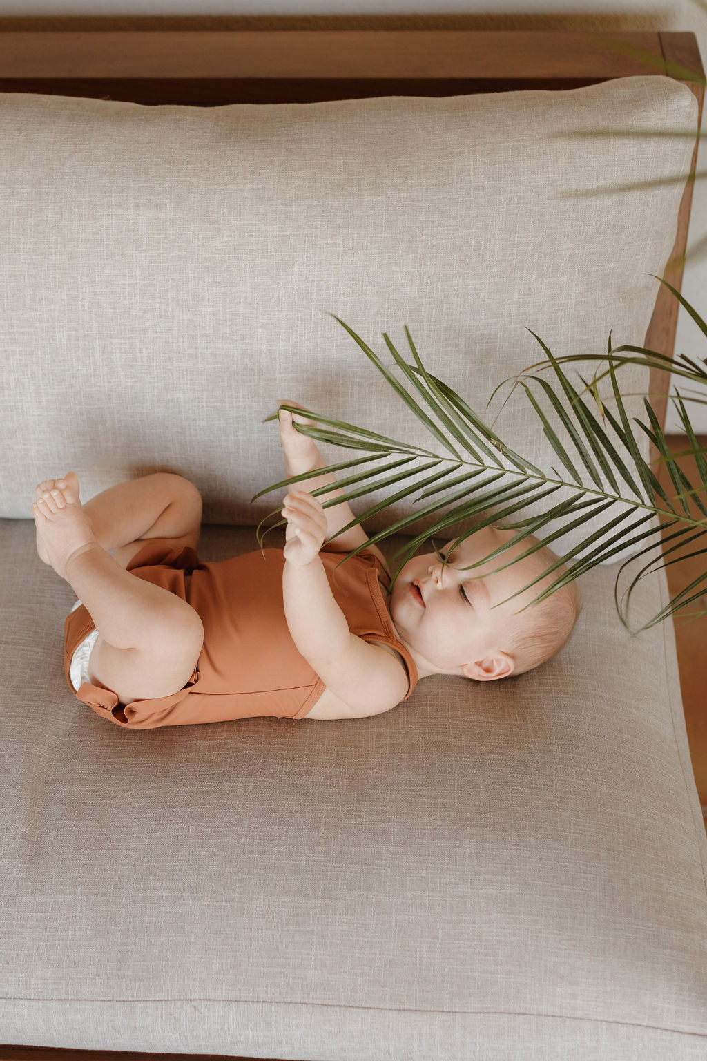 A baby dressed in a Girls Sleeveless Swimsuit from the Forever French Baby Terra Collection playfully lies on a light gray couch, holding a green palm frond. Soft lighting highlights the serene scene and the baby's peaceful expression.