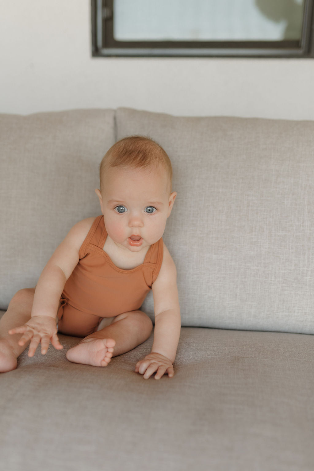 A baby in a Girls Sleeveless Swimsuit from the forever french baby Terra collection sits on a light gray couch with a curious expression, one hand outstretched and legs crossed, against a background of a white wall and framed window.