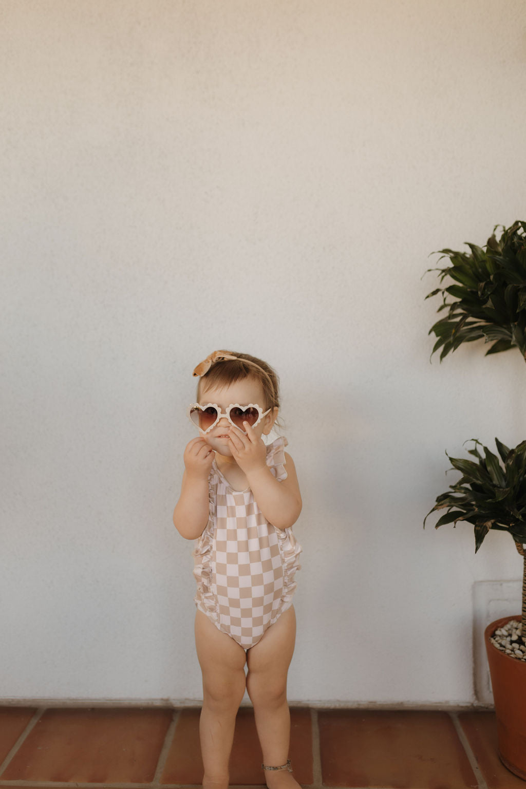 A toddler in a checkered "Girls Sleeveless Swimsuit | Coastline" from Forever French Baby and heart-shaped sunglasses stands indoors, adjusting their shades. On the right, a potted plant against a plain wall complements the timeless charm of this moment.