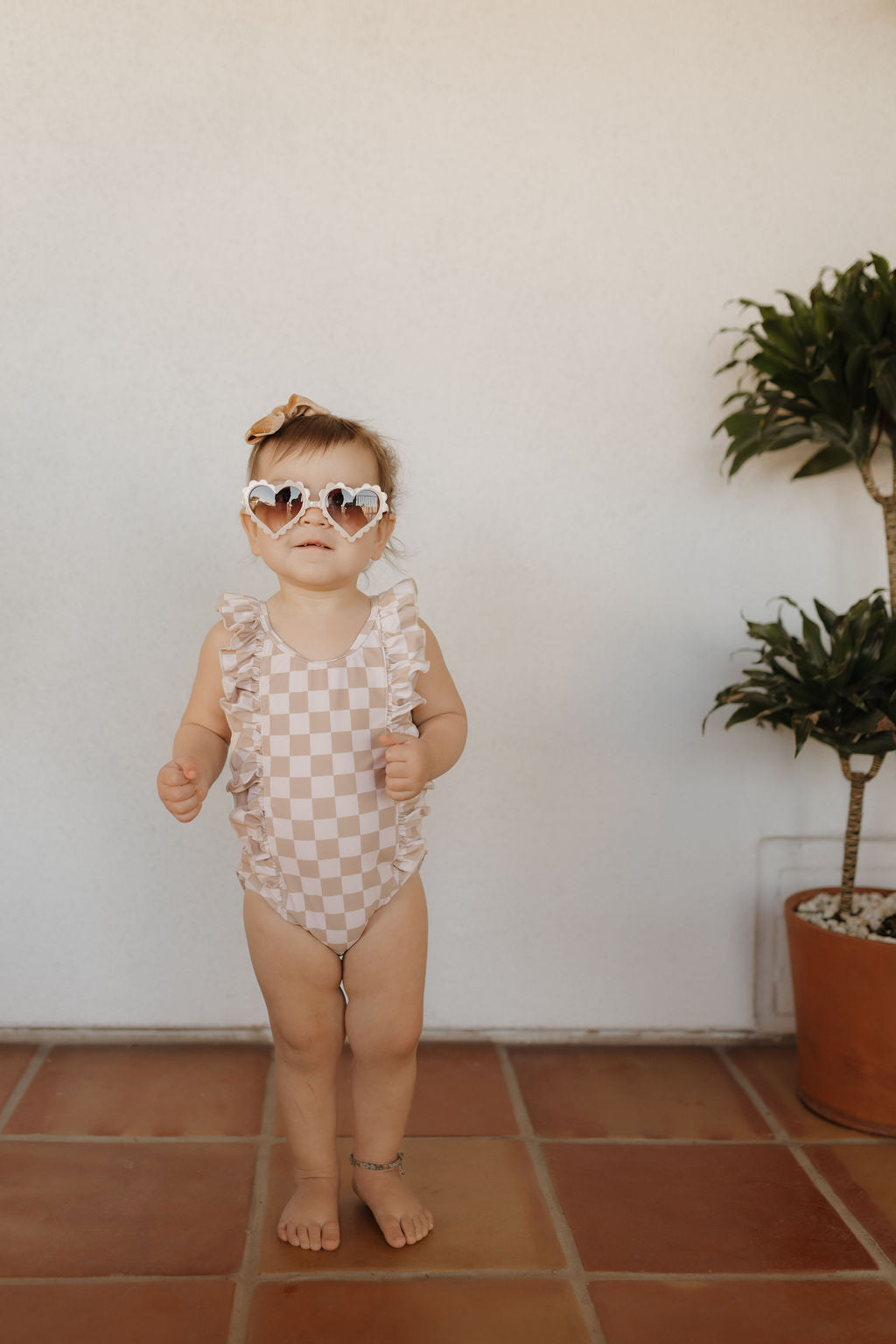 A toddler in a Girls Sleeveless Swimsuit from Forever French Baby's Coastline collection, heart-shaped sunglasses, and a hair bow stands barefoot on tiled flooring. Posed beside a potted plant with a plain white wall background, they embody the essence of Forever French Baby style.