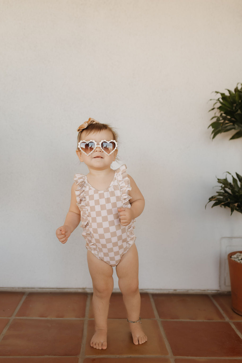 A toddler models forever French baby's Girls Sleeveless Swimsuit from the Coastline Collection on a tiled floor, accessorized with heart-shaped sunglasses, a small hair bow, and a delicate anklet. Potted plants enhance the charm against a plain wall.