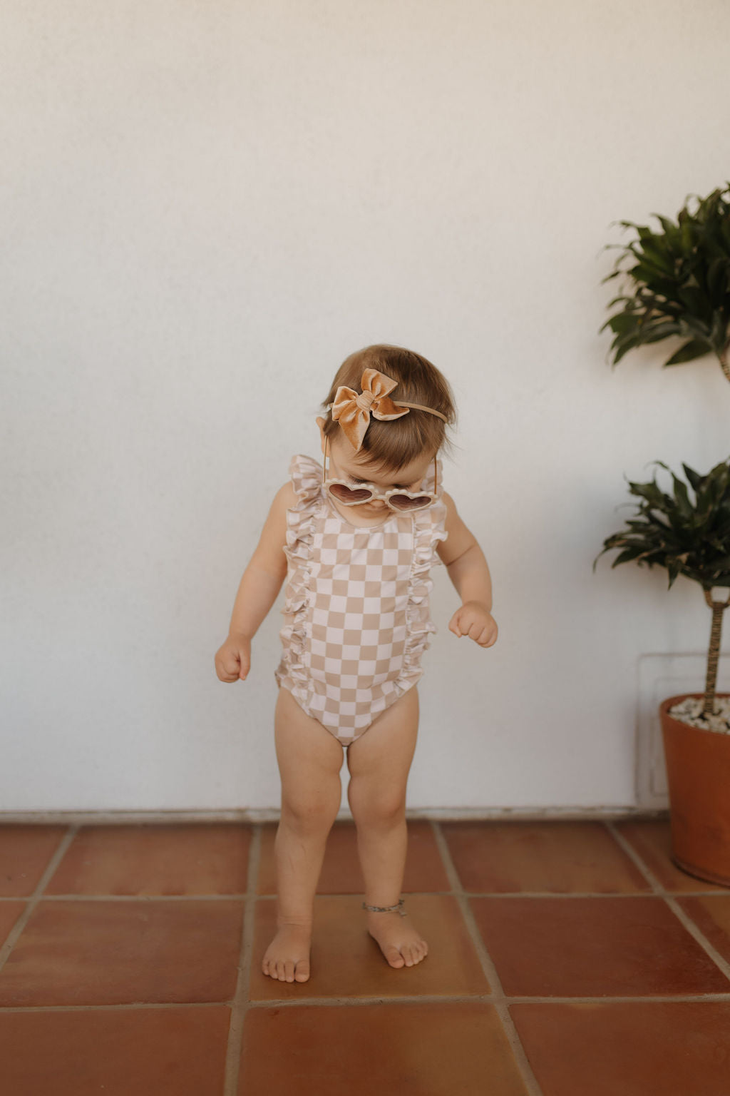 A toddler from forever French baby is on a tiled floor, wearing a timeless Girls Sleeveless Swimsuit from the Coastline Collection. She's accessorized with sunglasses and a small bow in her hair, standing gracefully next to a lush potted plant.