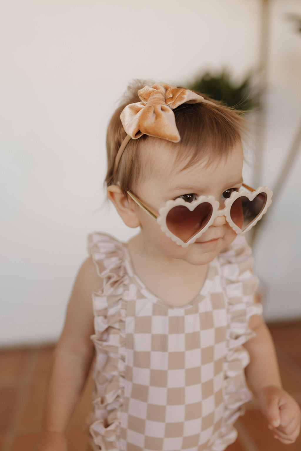 A toddler from the forever French baby Coastline collection wears a checkered sleeveless swimsuit, heart-shaped sunglasses, and a peach bow headband, standing on a tiled floor and gazing slightly to the side against a softly blurred background.