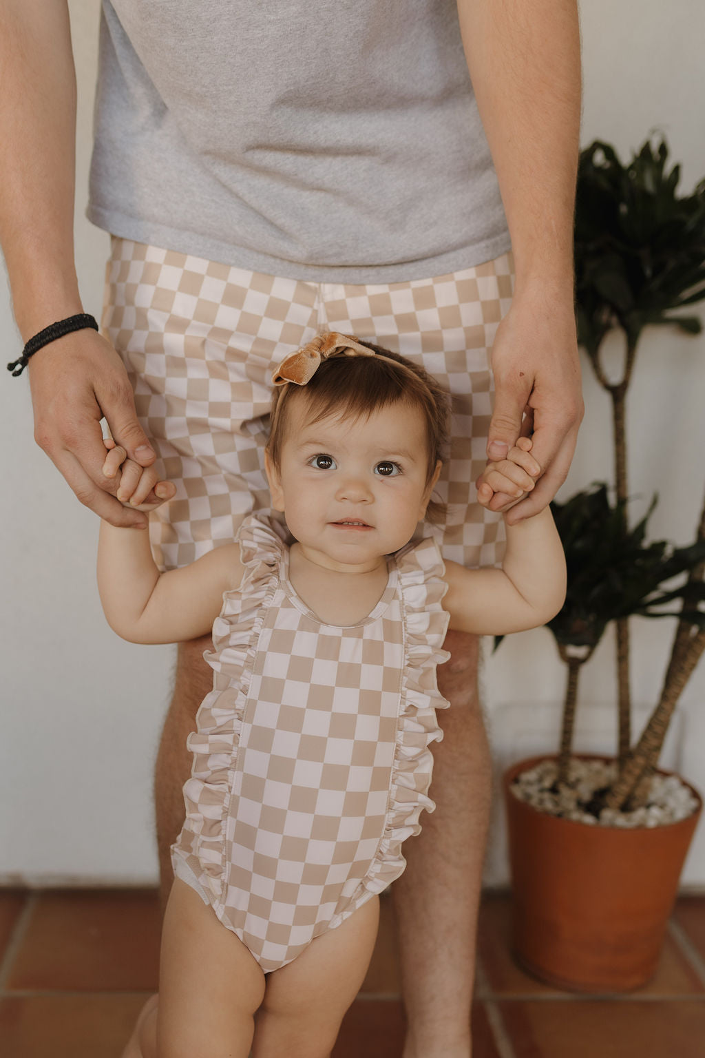 A toddler in a Forever French Baby Girls Sleeveless Swimsuit from the Coastline collection holds an adult's hands, both stylishly matched while a potted plant enhances the scene's charm.