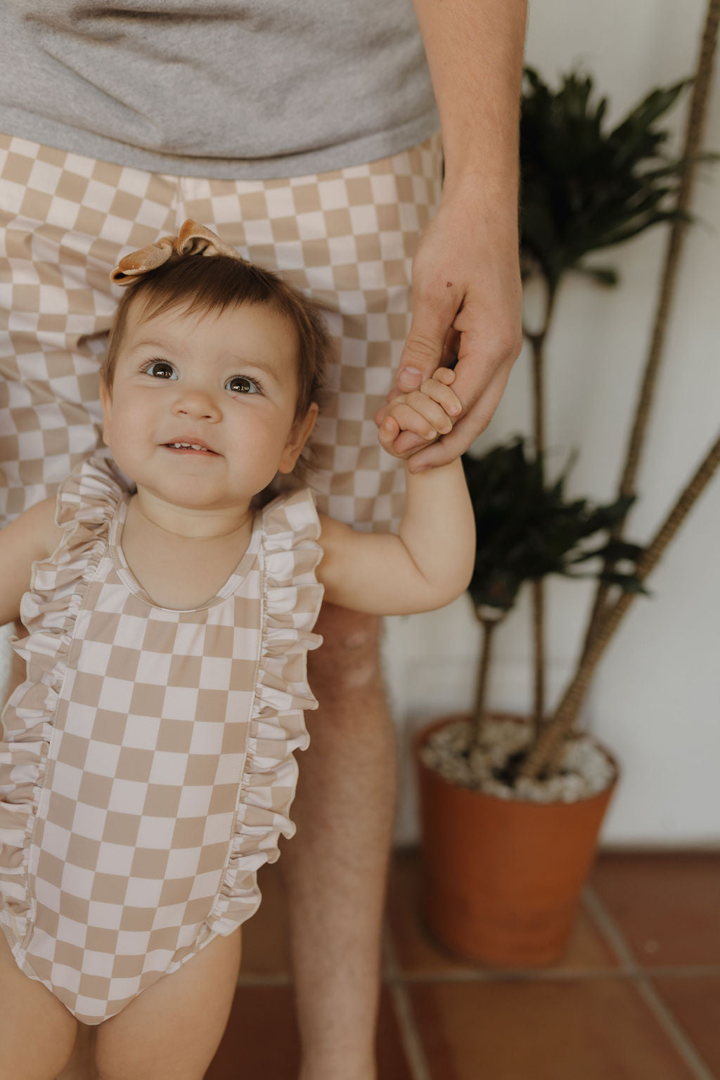 A baby in a checkered swimsuit holds hands with an adult in matching Men's Boardshorts from the Coastline line by forever french baby, standing on terracotta tiles with a potted plant behind them.