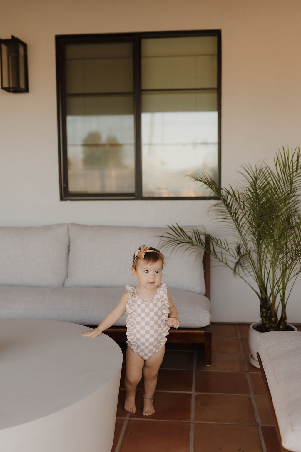 A toddler in a classic Girls Sleeveless Swimsuit from the Coastline collection by forever French baby stands near a round white patio table. A beige sofa and potted palm are behind, with a large black-framed window as the backdrop.