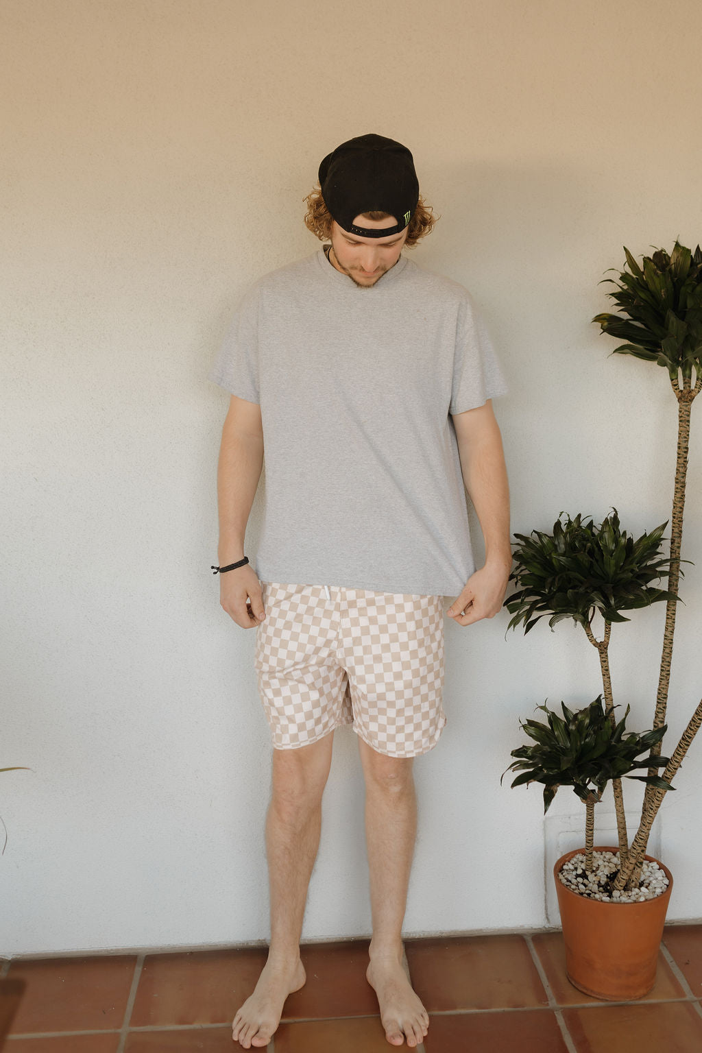 A person in a backward cap and gray shirt wears checkered Men's Boardshort | Coastline from forever french baby, standing on a tiled floor with potted plants nearby against a plain light-colored wall.