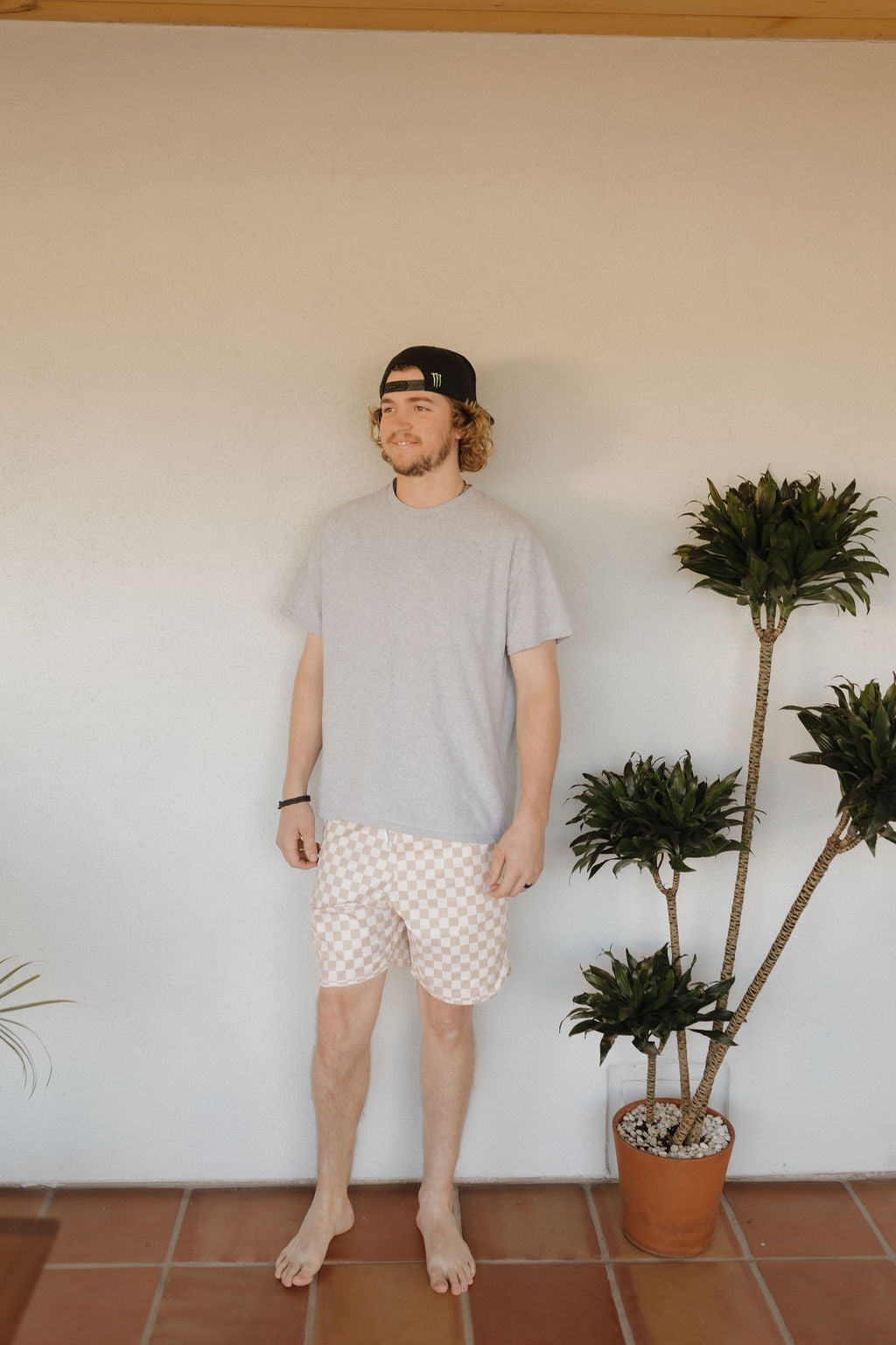 A curly-haired man in a black cap and Men's Boardshort from Coastline by forever french baby stands barefoot on a tiled floor, next to a tall potted plant against a plain wall, exuding relaxed vibes.