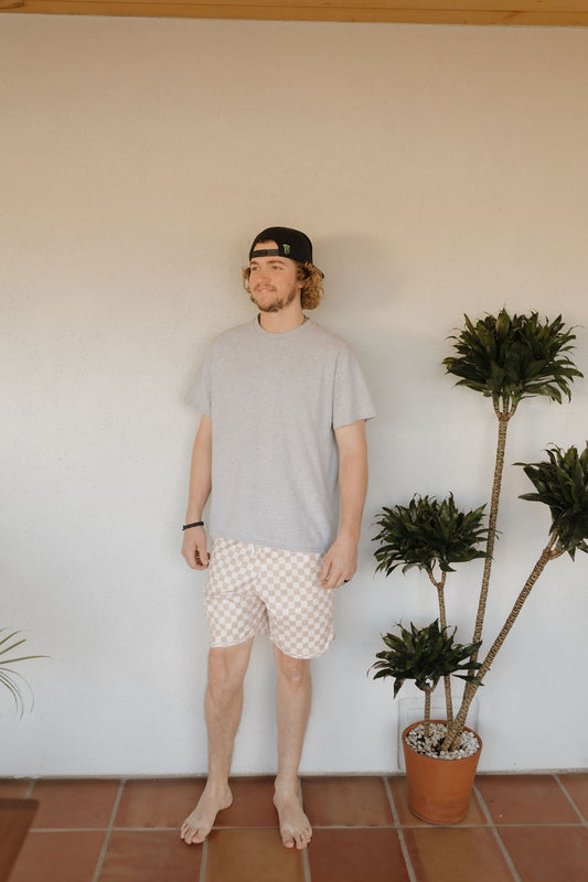 A curly-haired man in a black cap and Men's Boardshort from Coastline by forever french baby stands barefoot on a tiled floor, next to a tall potted plant against a plain wall, exuding relaxed vibes.