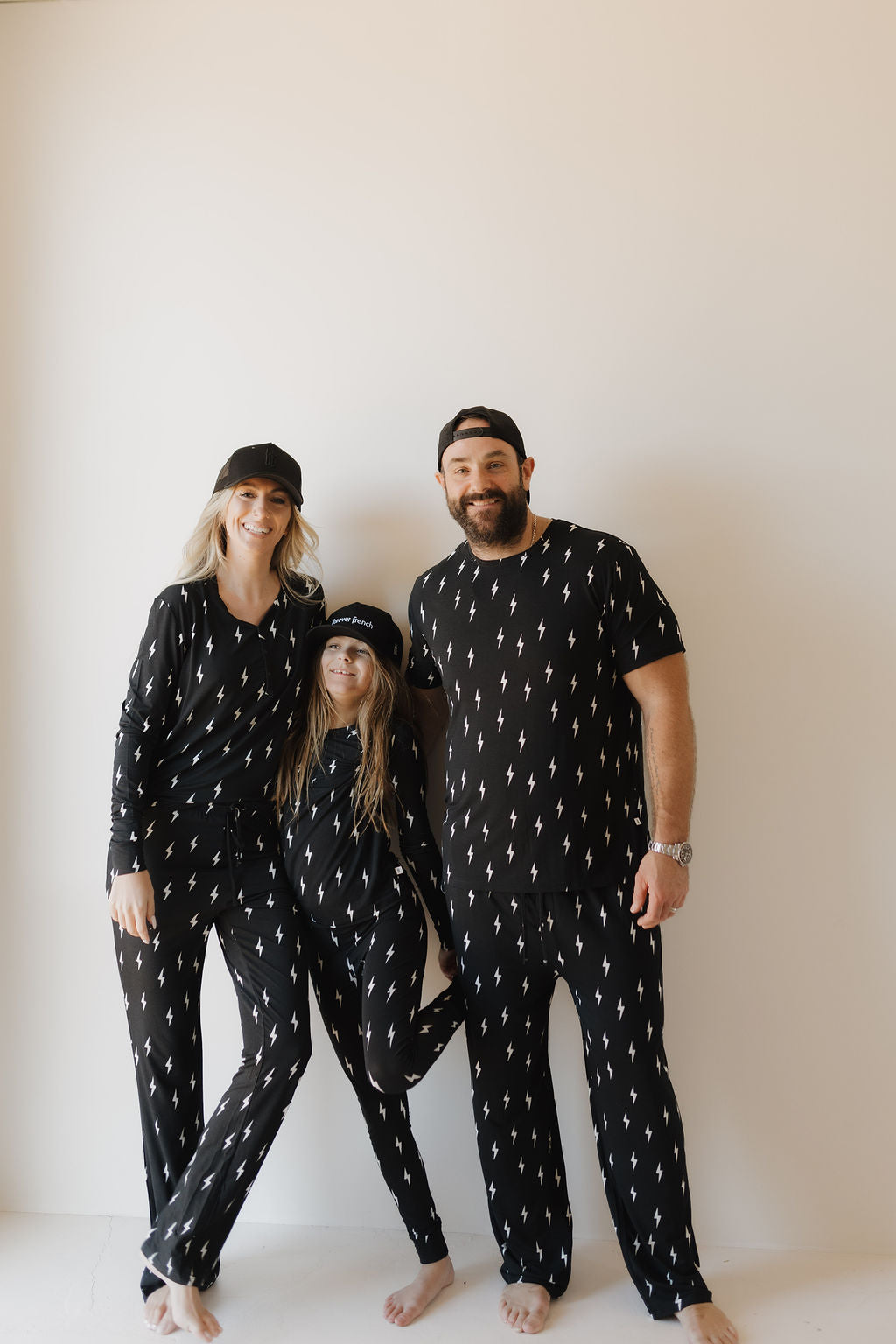 Against a plain backdrop, a smiling family of three showcases matching "Midnight Lightning Bolt" bamboo pajamas from forever french baby. The two adults flank the child, who wears a cap and leans on one of them.