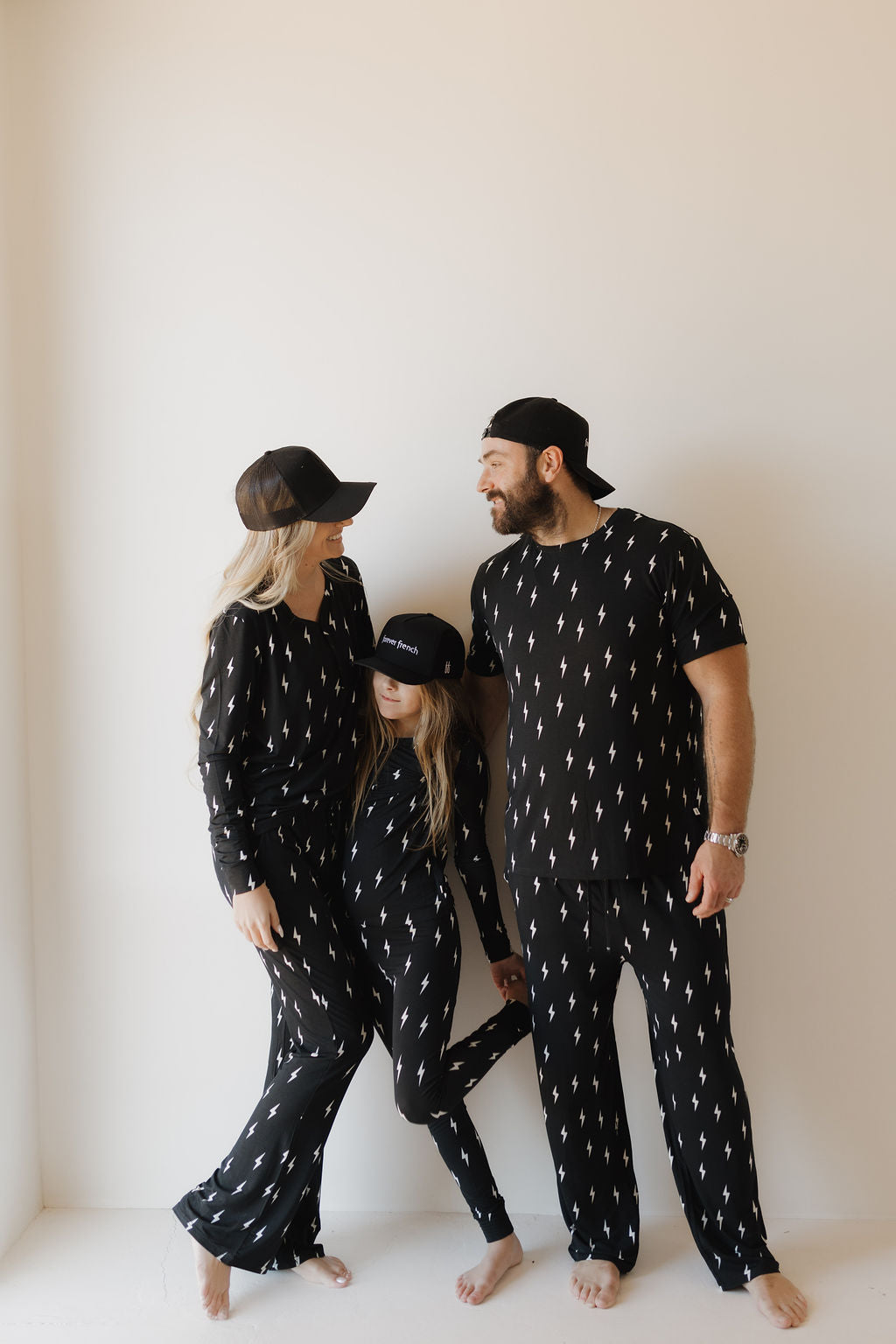 A family of three in matching "Midnight Lightning Bolt" bamboo pajamas and baseball caps by forever french baby stands barefoot in a corner, with the parents sharing a look and their child between them.