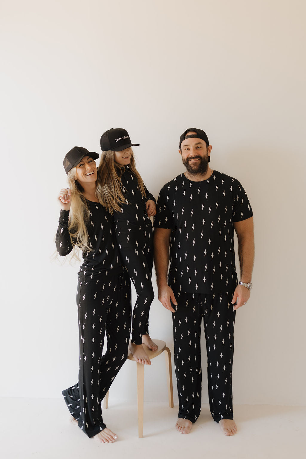 A family of three poses in matching "Bamboo Two Piece Pajamas | Midnight Lightning Bolt" by forever french baby, featuring a small white lightning bolt pattern. The woman and girl wear black caps, while the man smiles. The little girl stands on a stool against a light background, loving the breathable comfort.