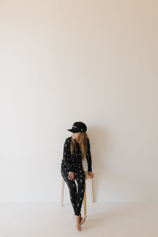 A person wearing Bamboo Two Piece Pajamas in Midnight Lightning Bolt by forever french baby and a cap, resembling breathable sleepwear, sits barefoot on a stool against a plain white background, looking to the side.