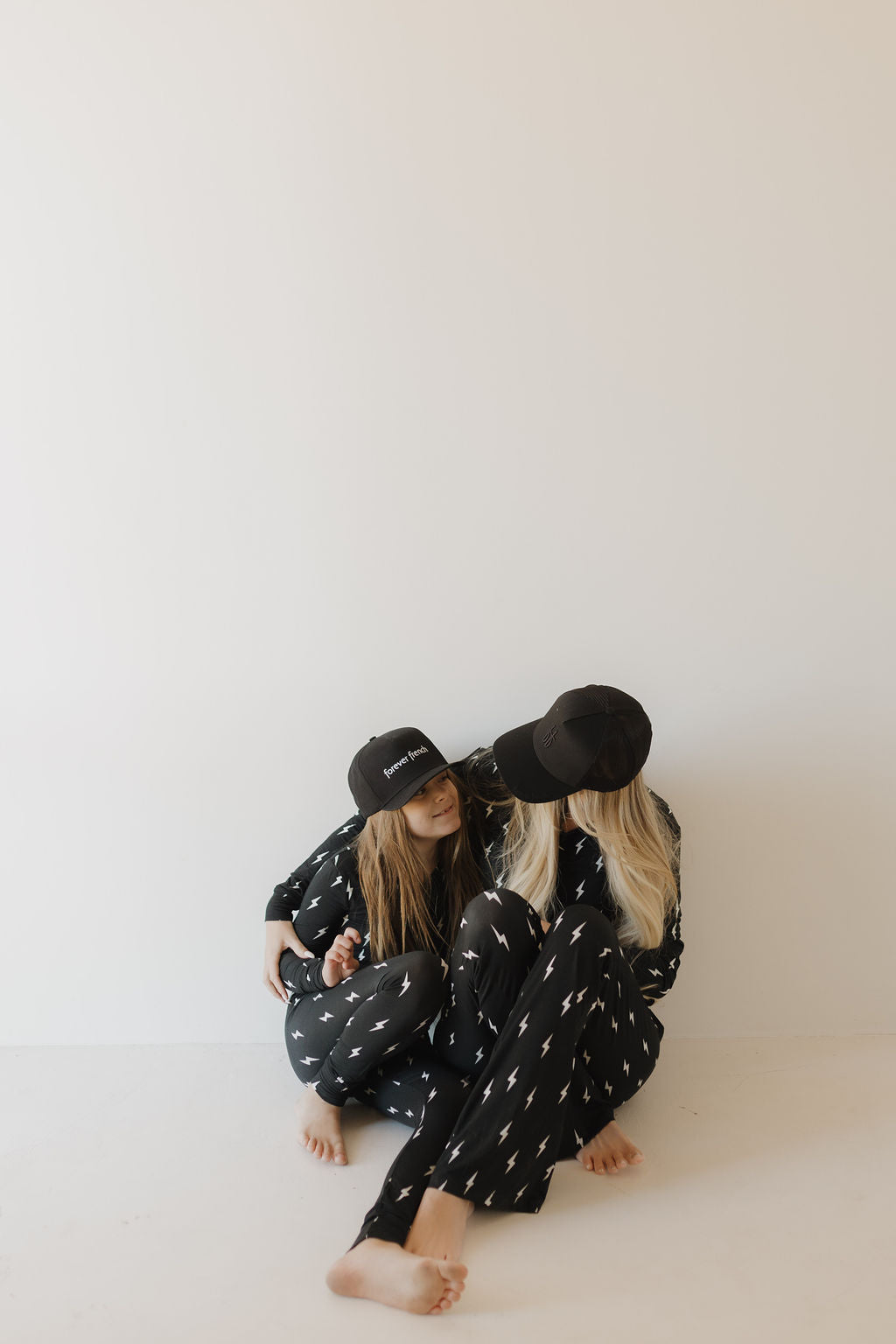 Two people sit on the floor against a white wall, both wearing forever french baby Women's Bamboo Pajamas in the Midnight Lightning Bolt pattern and matching black caps. The breathable fabric enhances the cozy and relaxed setting as they interact with each other.