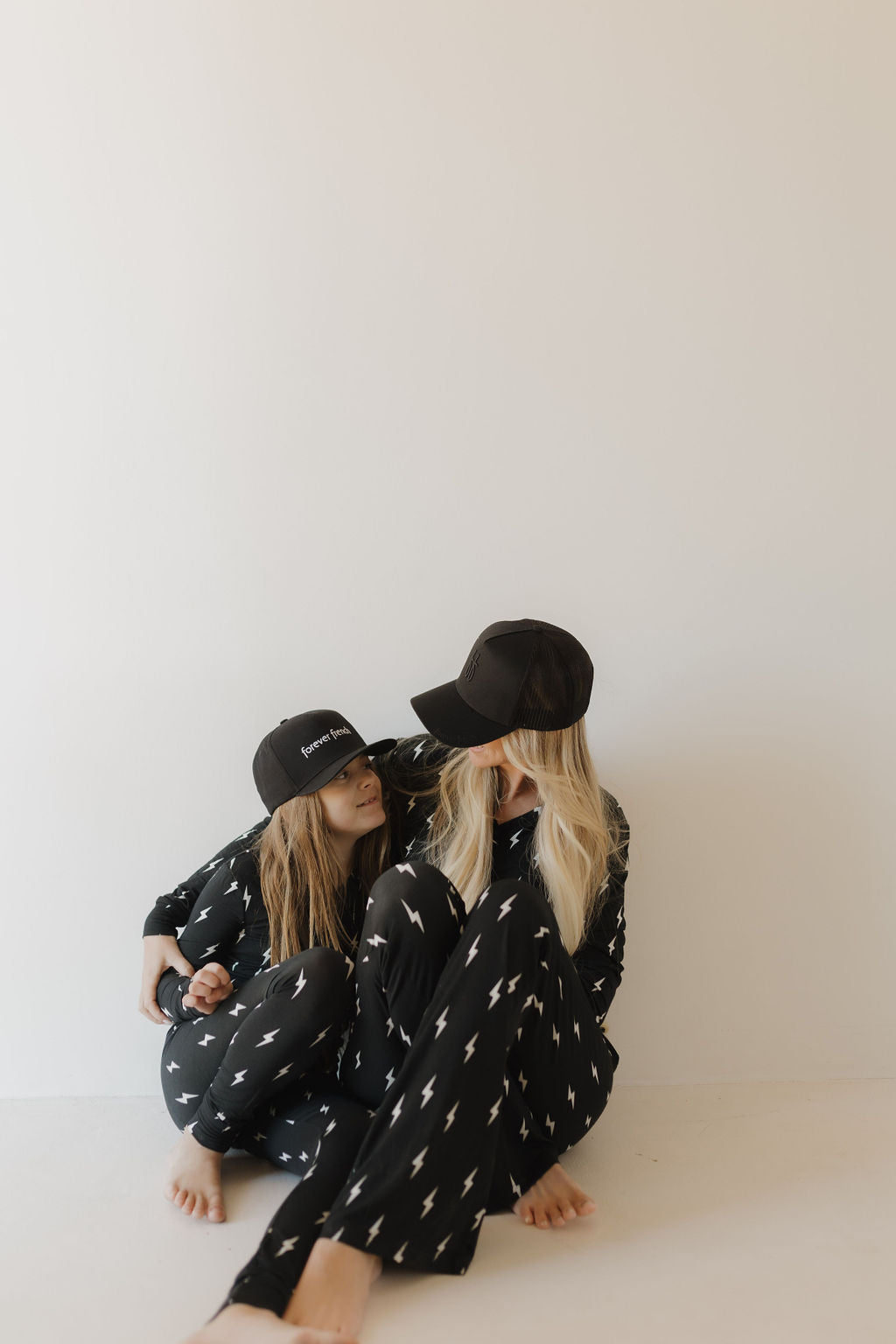 Two individuals sit on the floor against a plain background in matching forever french baby Bamboo Two Piece Pajamas | Midnight Lightning Bolt, featuring black outfits with white lightning bolt patterns and black hats. The breathable sleepwear offers comfort as one leans on the other's shoulder.