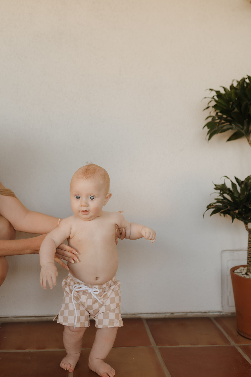 A light-skinned baby with short hair stands in Child Boardshort | Coastline by forever french baby on a terracotta tile floor. Gentle arms hold the baby, capturing the Forever French Baby vibe. A lush potted plant decorates the scene against a pristine white wall.