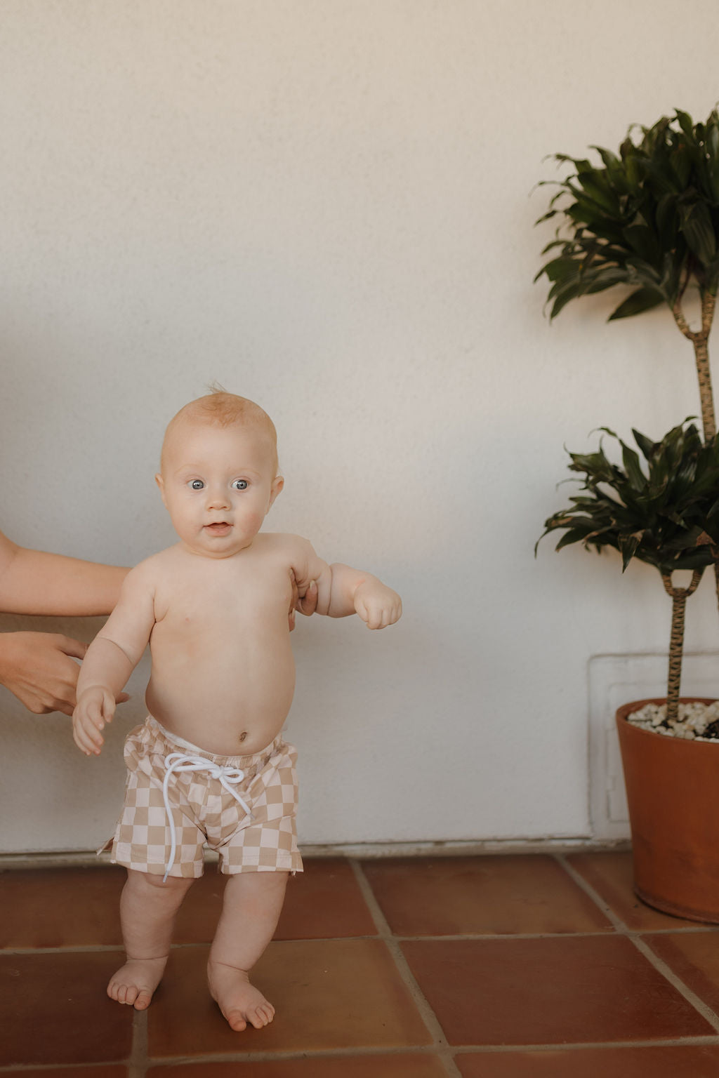 A baby in forever french baby's Child Boardshort | Coastline stands on a tiled floor, supported by an adult's hands. A potted plant sits on the right against an off-white wall, completing this charming scene.