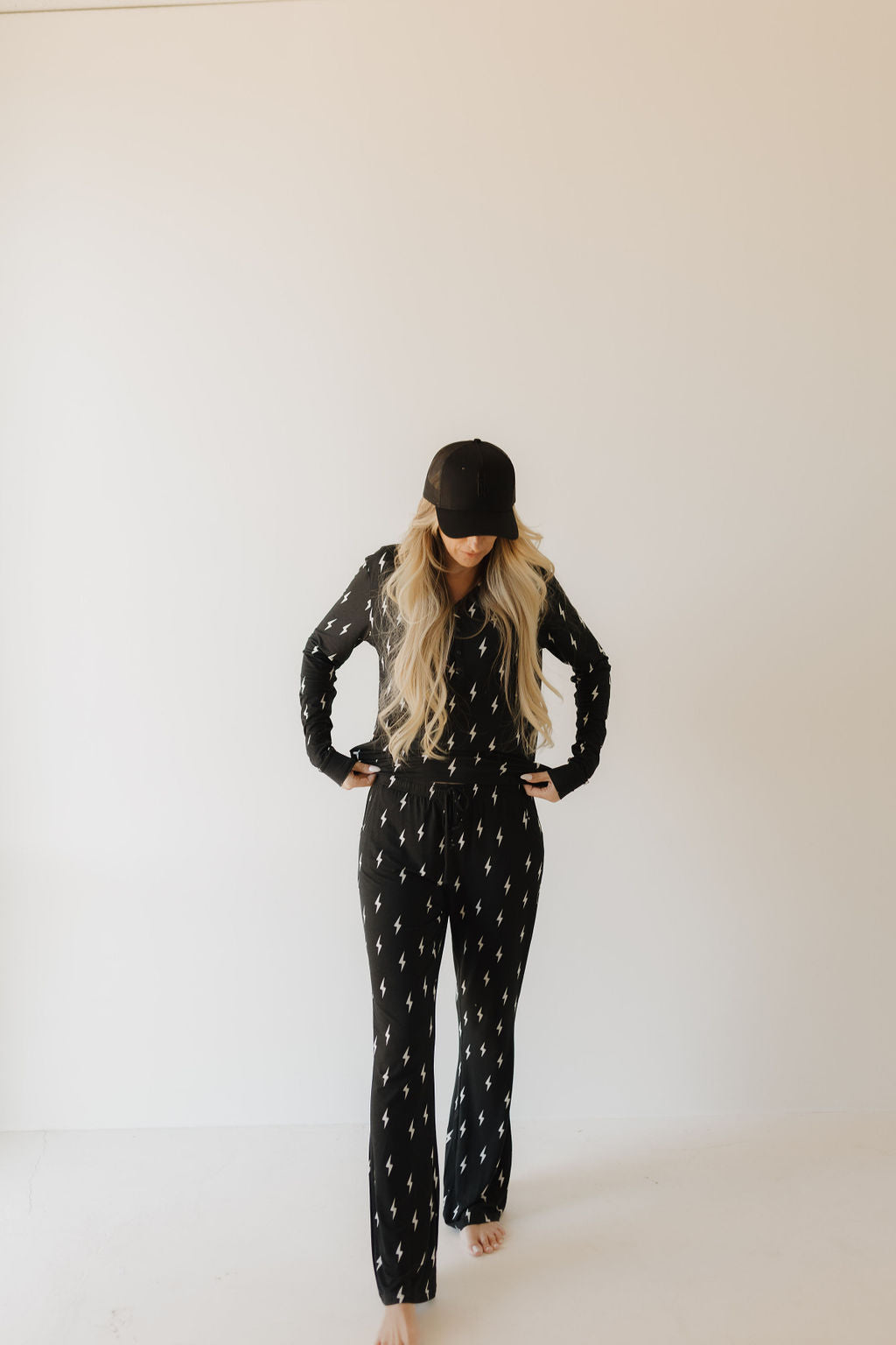 Wearing forever french baby's Women's Bamboo Pajamas in Midnight Lightning Bolt, a person poses against a plain background. Their black cap completes the look, while their hands on hips show off long, wavy hair.