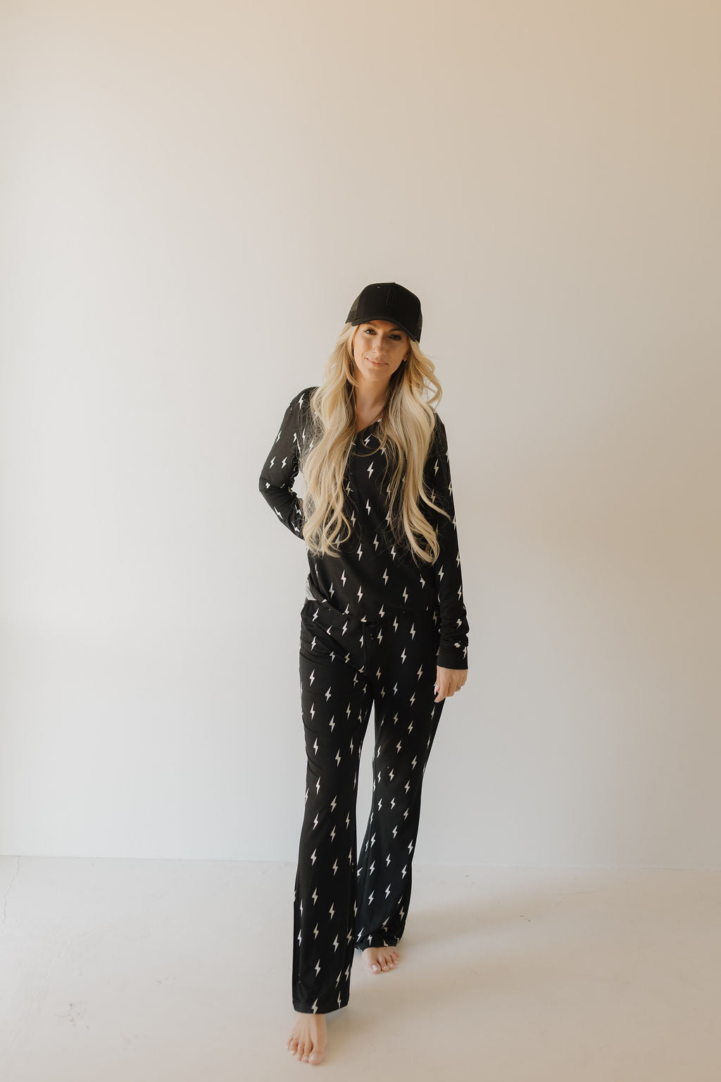 A person with long blonde hair wears a black hat and Women's Bamboo Pajamas in Midnight Lightning Bolt by forever french baby, featuring small white patterns. The breathable fabric matches their relaxed demeanor as they stand barefoot against a plain light background, smiling softly.