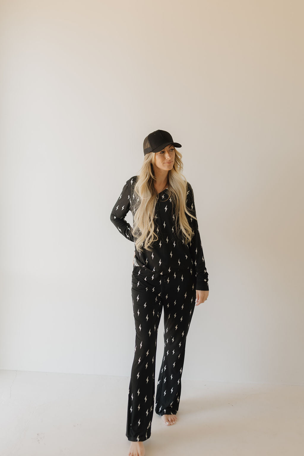 A woman wearing forever french baby's Women's Bamboo Pajamas in "Midnight Lightning Bolt," complemented by a black hat, stands barefoot against a plain light-colored background.