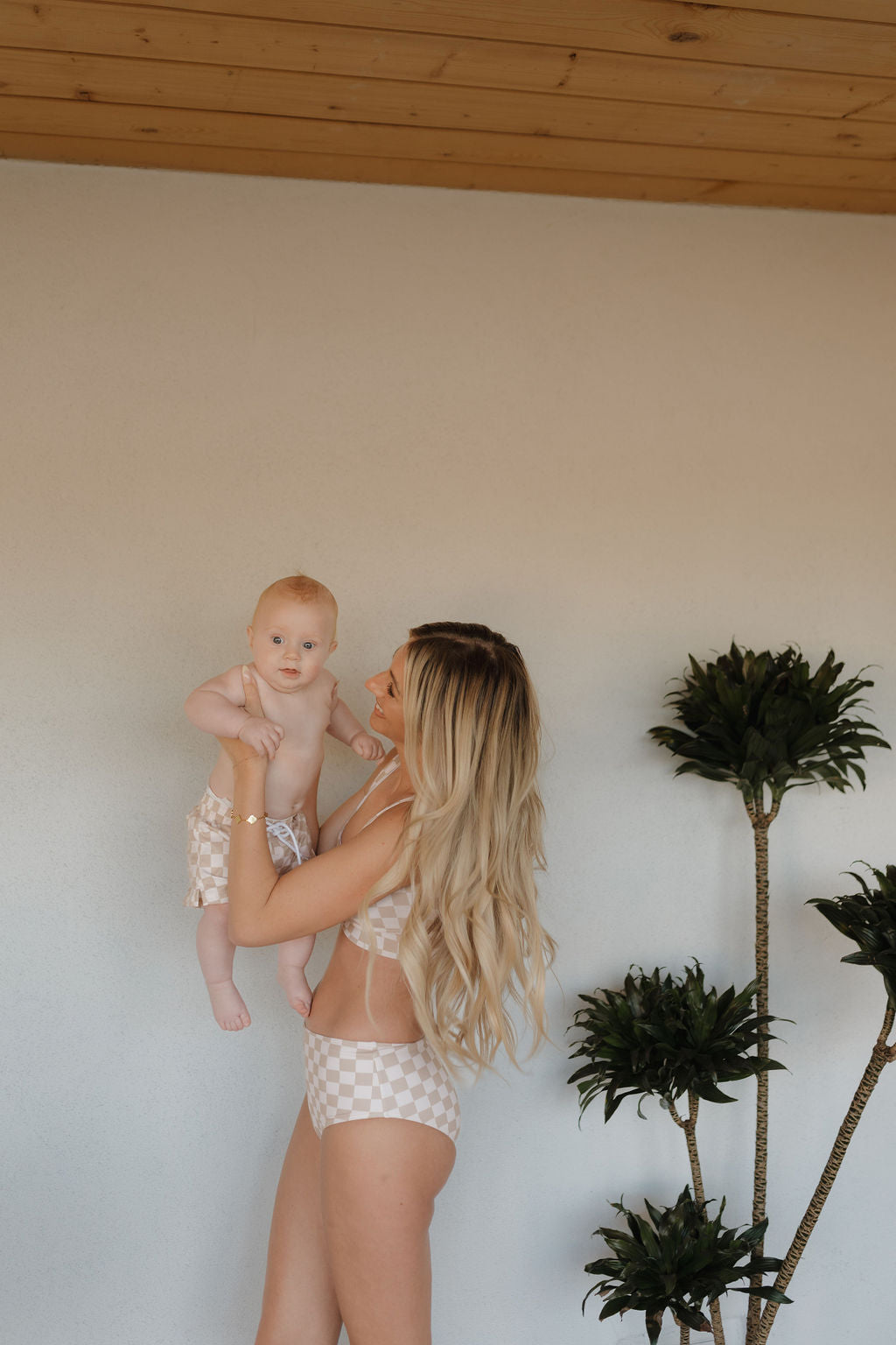 A woman with long, blonde hair cradles a baby in her arms. They sport checkered swimsuits from forever french baby's Coastline collection. Beside them stand two tall plants with slender trunks and green leaves against a light-colored wall.