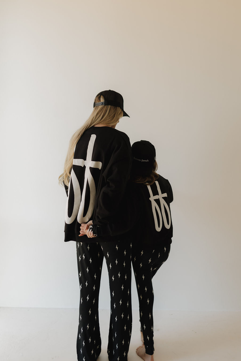 Two people of different heights stand back-to-back. Both wear matching black "Adult Sweatshirt | ff Black" by forever french baby, paired with black pants and caps, all 100% cotton. The garments feature white symbols and patterns. The background is plain and light-colored.