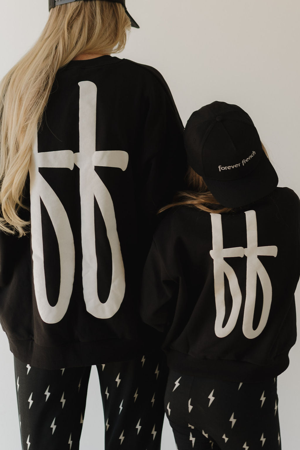 Two people wear outfits with bold logos. The person on the left wears the "Snapback | forever french Black" by forever french baby, their long hair cascading down. The shorter individual also sports a cap. Both are turned away, emphasizing their attire's striking logo stitching.