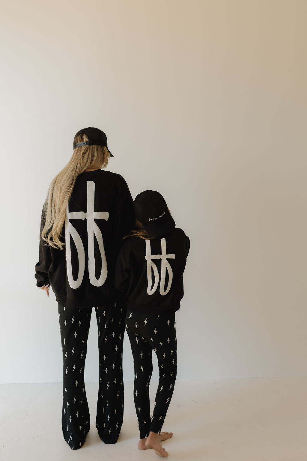 Two people stand facing away from the camera in black clothing, each adorned with the bold FF logo design. The taller wears a cap; the shorter has a hooded jacket and cap. Their matching patterned pants are 100% cotton. The backdrop is plain, featuring Forever French Baby's Adult Sweatshirt | ff Black.