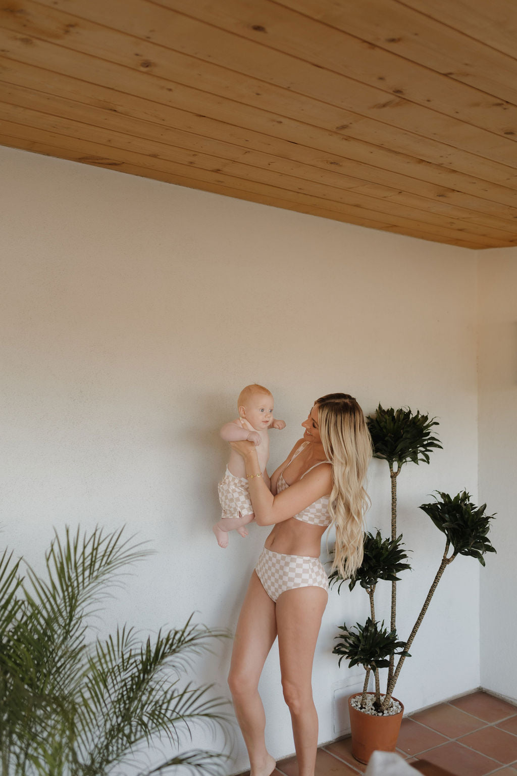 A blonde woman cradles a baby, both in matching checkered swimsuits from the Coastline collection by Forever French Baby, standing on a tiled floor near potted plants under a wooden ceiling.