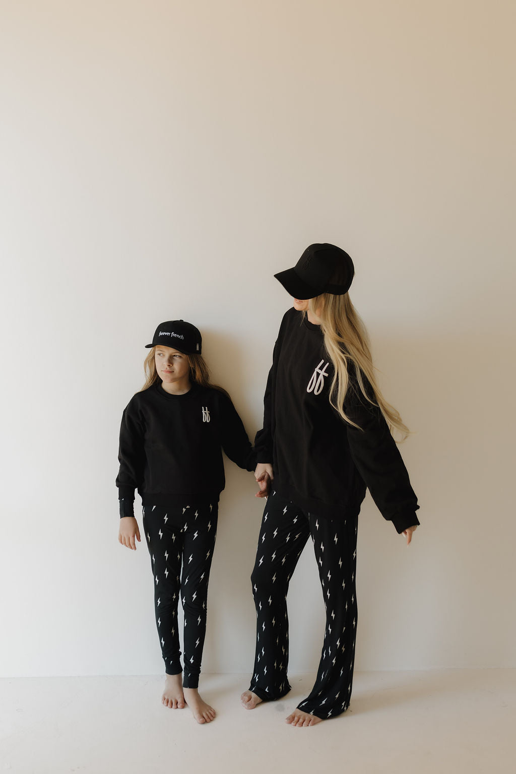 A barefoot woman and child sport matching "ff Black" sweatshirts from forever french baby. Their black outfits adorned with white designs add flair as they stand hand-in-hand against a light wall, facing slightly away from the camera.