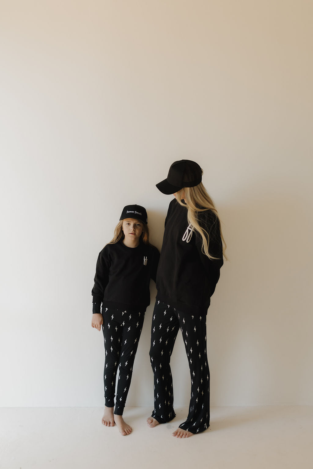 Two people, one taller and one shorter, both wearing black Adult Sweatshirts by forever french baby featuring the iconic FF logo, stand barefoot in black caps against an off-white wall, gazing to the side. Their cozy matching outfits are crafted from 100% cotton.