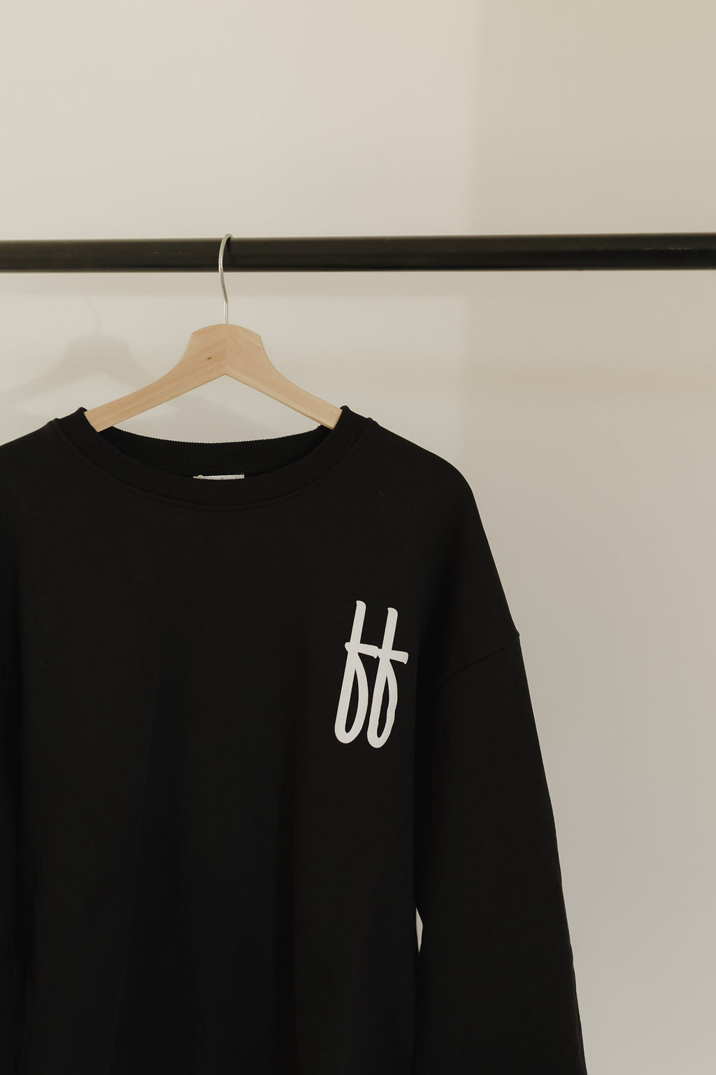 The Child Sweatshirt | ff Black by forever french baby, crafted from 100% cotton, showcases a white "ff" logo on the left side. It hangs stylishly on a wooden hanger against a plain white wall, displayed on a sleek black rail rod.