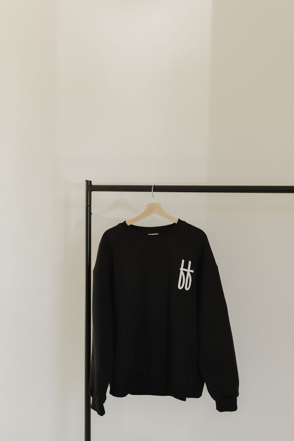 The Child Sweatshirt | ff Black by forever french baby, a cozy 100% cotton black sweatshirt with a white FF logo, hangs invitingly on a wooden hanger on a sleek black metal clothing rack against a plain white wall.