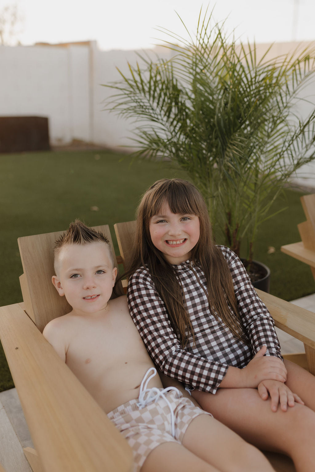 Two kids are sitting on a wooden chair outdoors, both smiling at the camera with a green plant behind them. The child on the left wears checked shorts similar to Child Boardshort | Coastline, and the one on the right dons a long-sleeve checkered outfit from forever french baby's Coastline collection.