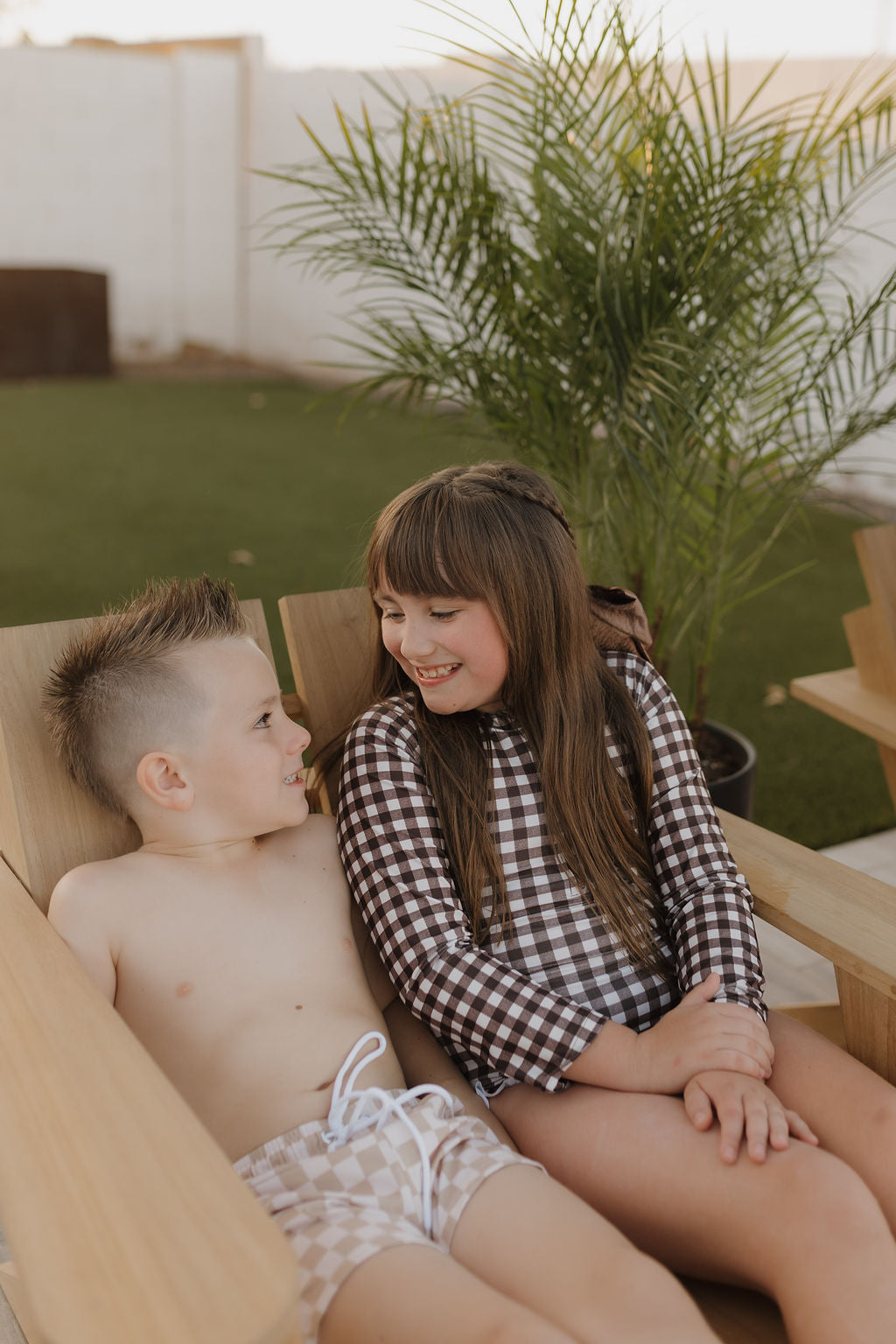 A boy and a girl are sitting on a wooden chair outside. The shirtless boy wears Coastline's Child Boardshort, while the girl shines in her Forever French Baby checkered dress. Both smile at each other, with a potted plant in the background.