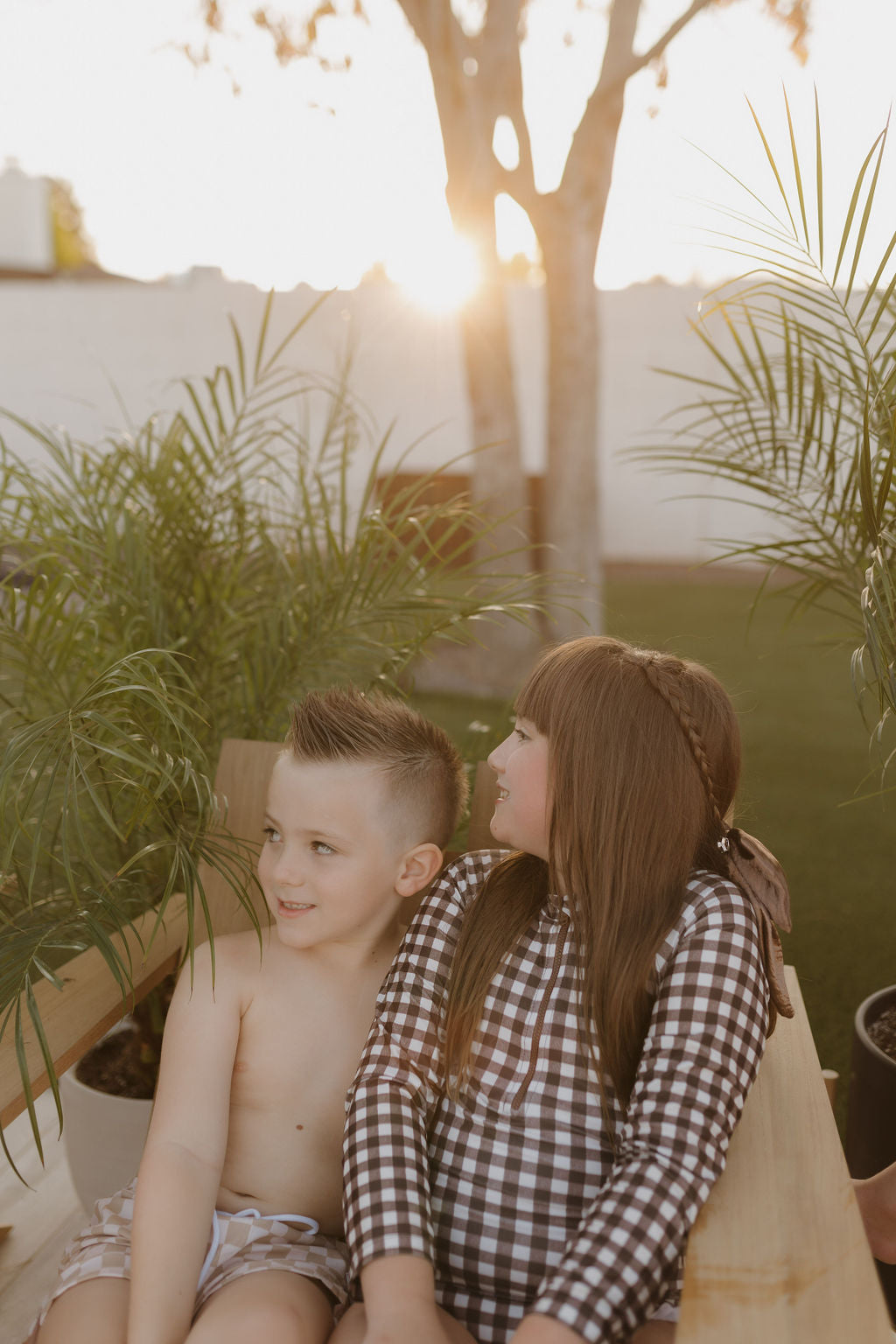 Two children sit together on a wooden bench outdoors, surrounded by potted plants. The sun sets, casting a warm glow. The shirtless child is beside another wearing a checkered Girls Long Sleeve Swimsuit from forever French baby's Chocolate Gingham Collection.