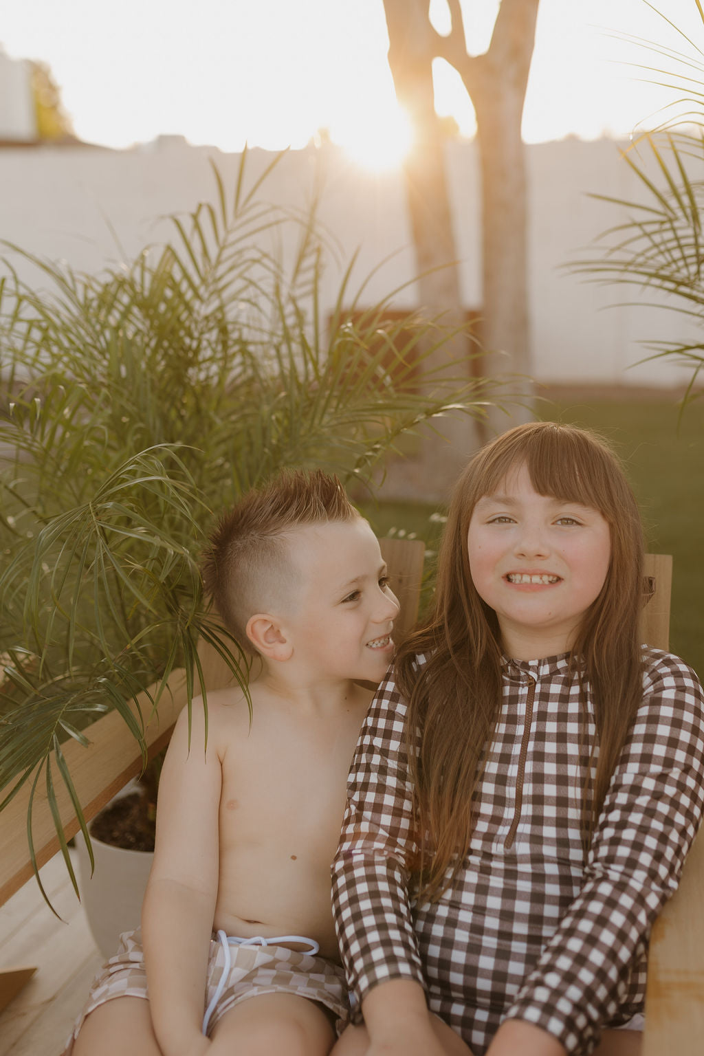 Two children sit together outside on a wooden bench. The shirtless boy, in vibrant Child Boardshorts from the forever french baby Coastline collection, has spiky hair and playfully looks at the girl. She's wearing a checkered outfit from the same collection, smiling softly as sunlight filters through the trees.