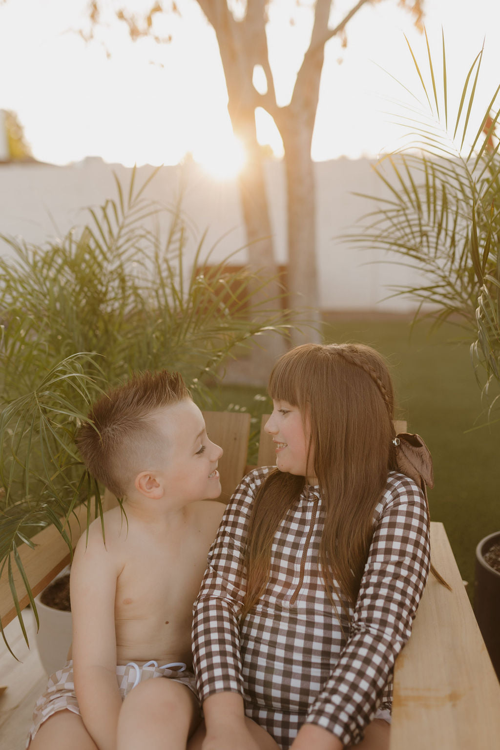 Two children sit on a wooden bench outdoors, surrounded by green plants. The boy wears Child Boardshort | Coastline, and the girl is in an outfit from the Forever French Baby Coastline collection. They smile at each other as the sun sets, casting a warm glow.