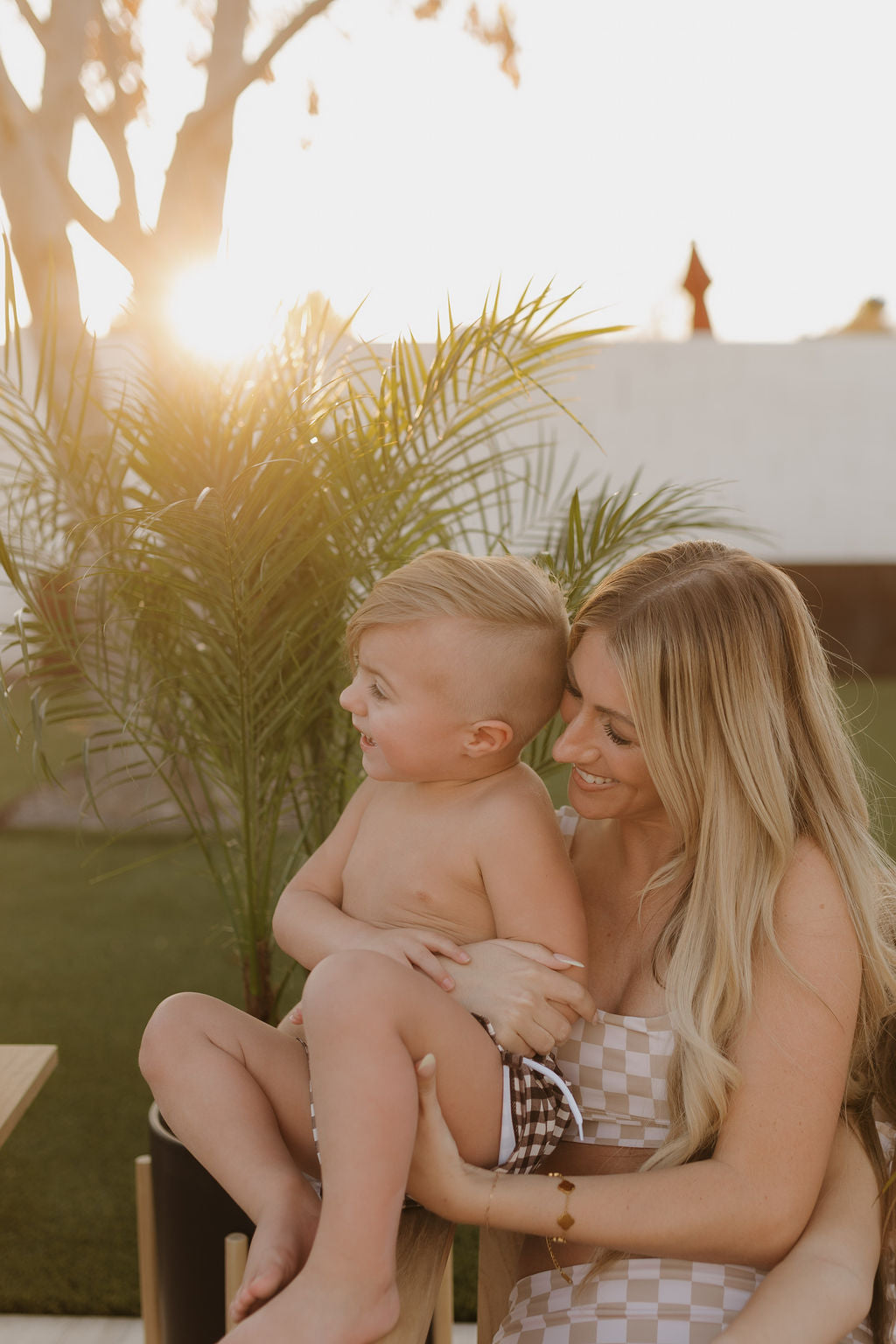 A woman with long blonde hair, wearing a Women's Two Piece Swimsuit from the Coastline collection by forever french baby, smiles while holding a laughing, shirtless child in checkered shorts. They are outdoors against a palm backdrop and stunning sunset.