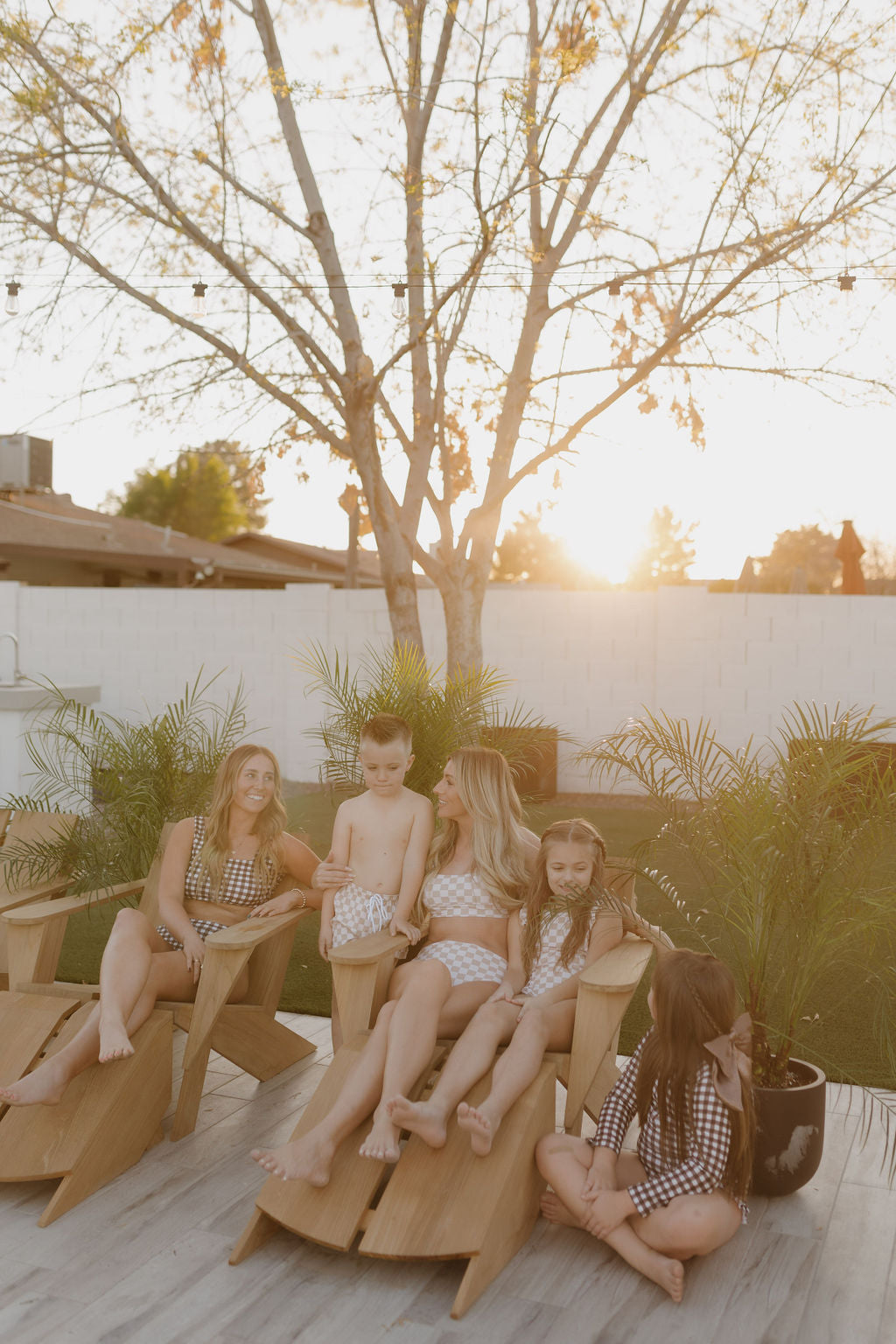 A family, dressed in timeless Girls Sleeveless Swimsuits from the Coastline collection by forever French baby, relaxes on wooden lounge chairs outdoors at sunset. Surrounded by small potted plants, they converse against a backdrop of a leafless tree and white brick wall.