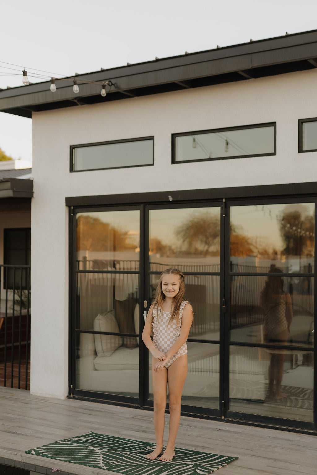 A young girl in a forever French baby Girls Sleeveless Swimsuit from the Coastline collection stands on a deck before a modern house with large glass doors. She smiles on a green and white mat, while string lights twinkle above her.